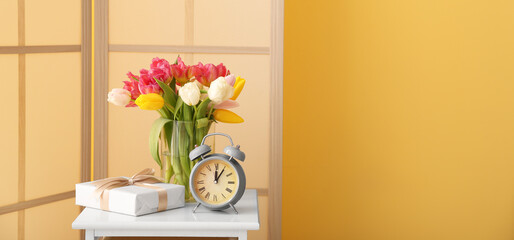Vase with beautiful tulip flowers, alarm clock and gift on table in room. Banner for design