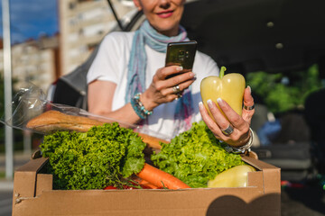 Fototapeta premium One woman mature caucasian female sit in the back trunk of her car on the parking lot of the supermarket shopping mall or grocery store with vegetables food in box putting them in the vehicle