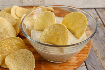 Board with delicious potato chips and bowl of sauce on grey wooden background