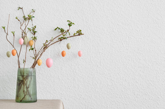 Vase with tree branches and Easter eggs on table near light wall