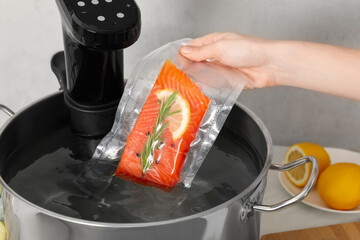 Woman putting vacuum packed salmon into pot with sous vide cooker in kitchen, closeup. Thermal...
