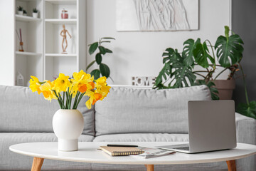 Modern laptop, vase with narcissus flowers, notebook and magazine on coffee table in living room