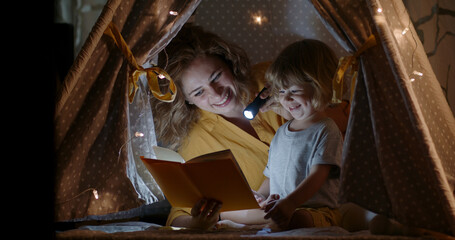 Obraz na płótnie Canvas Beautiful caucasian woman taking care of her child. Young mom is reading book of fairy tales for kid. Family in cozy tent in bedroom in evening 