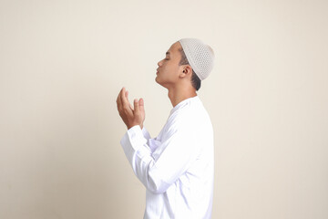 Portrait of attractive Asian muslim man in white shirt with skullcap praying earnestly with his hands raised. Isolated image on gray background