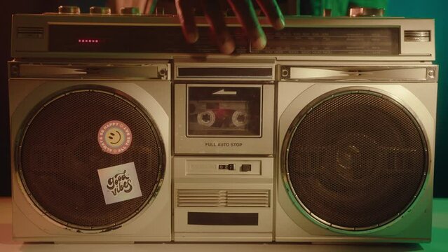 Close-up shot of hand putting compact audio cassette in ghetto blaster, pressing turn on button and playing the music in the studio with neon light