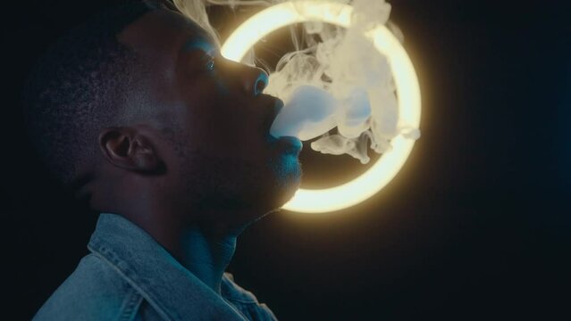 Slow motion footage of African-American man exhaling out vape smoke against glowing ring light in a dark studio. Side view medium close-up shot