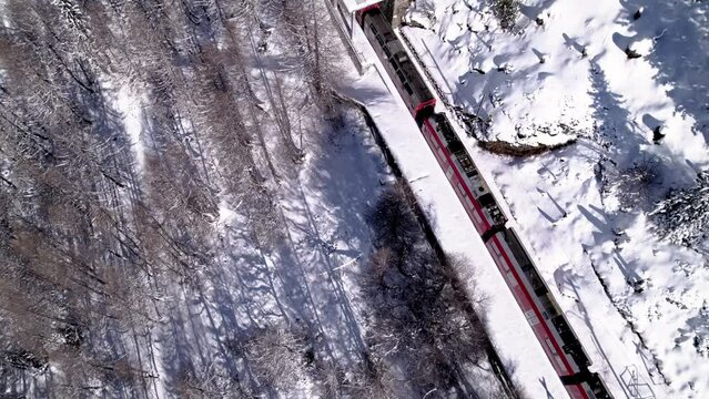 Aerial view of the Bernina Express panorama train going through a snow covered mountain winter landscape with forests on a sunny day in Alp Grum, Switzerland.