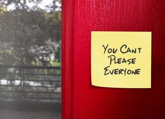 Yellow note stick on red wall with handwritten text You Can’t Please Everyone - self reminder to...