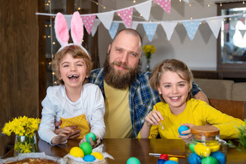 Easter Family traditions. Father and two caucasian happy children with bunny ears dye and decorate eggs with paints for holidays while sitting together at home table. Kids embrace and smile in cozy