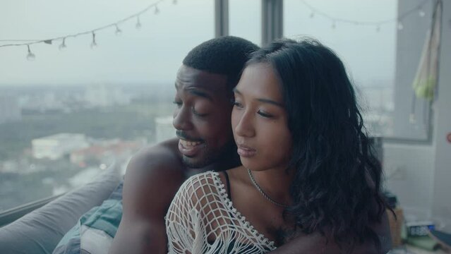 Young black boyfriend hugging his beautiful girlfriend when sitting together by the window overlooking cityscape, speaking and having romantic time at home