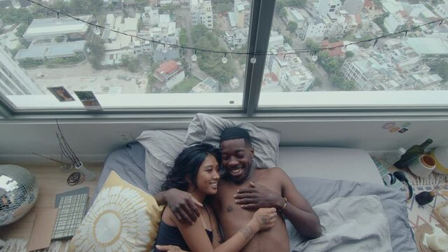 Young loving couple embracing, smiling and talking when lying in bed after waking up in the morning. Cityscape is seen from the apartment window. Top down shot