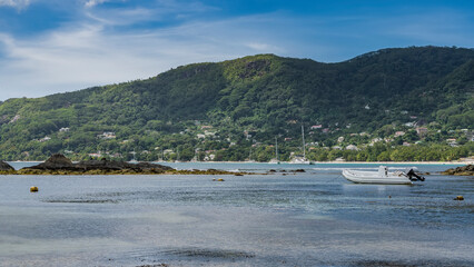 Obraz premium The rocky seabed was exposed at low tide. The boat is moored in shallow water. Yachts in the distance. A green hilly island against a background of blue sky and clouds. Seychelles. Mahe