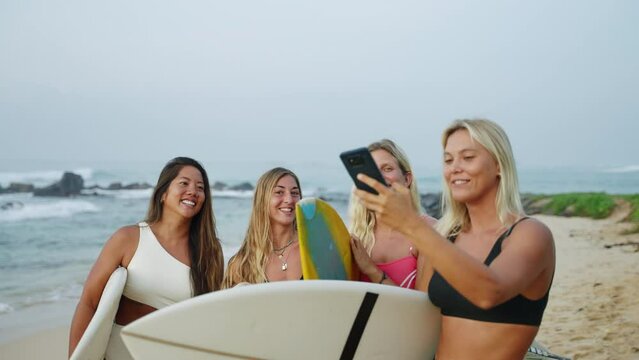 Cheerful multiracial surfer girls in bikini taking selfies holding surfboards on beach then look at phone screen. Sexy beautiful young surfing women in swimwear take selfie photographs after session.