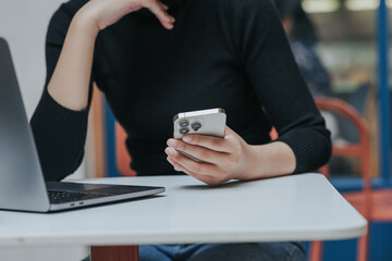 Young Asian woman using smartphone and laptop working remotely at coffee shop.