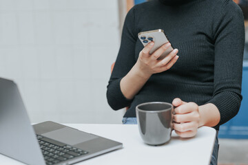 Young Asian woman using smartphone and laptop working remotely at coffee shop.