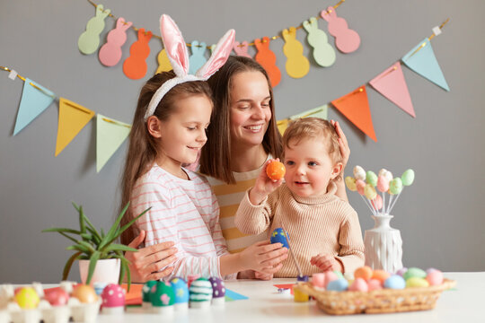 Indoor shot of mother and daughters painting eggs together preparing for Easter against gray decorated wall, infant baby holding colored egg, family spending time together.
