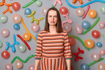 Obraz na płótnie Canvas Portrait of sad despair woman with brown hair wearing striped dress standing against gray wall with colorful balloons, looking at camera with sadness, nobody come to her birthday party.