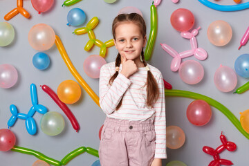 Image of cute charming smiling little girl with braids wearing casual clothing posing isolated over gray background with balloons, holding her chin, showing thumb up.