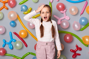 Photo of shocked amazed little girl with braids wearing casual clothing standing against gray wall with colorful balloons, keeps hand on forehead, forgot.