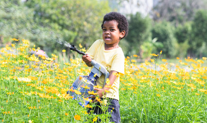 African 3 years old boy holding a big and heavy watering can, watering flowers garden on sunny day. Cute little child gardening, spraying water from a watering can at backyard in summer. Funny moment © Pruksachat