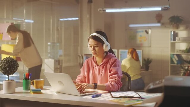 A young woman in white large headphones is typing on a laptop keyboard and enjoying music. In the background, a young creative team of managers in bright sweatshirts work.