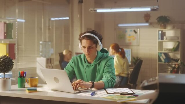 A young man in white large headphones is typing on a laptop keyboard and enjoying music. In the background, a young creative team of managers in bright sweatshirts work.