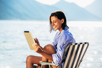 Smiling woman sitting on deck chair by the sea using digital tablet on a sunny day