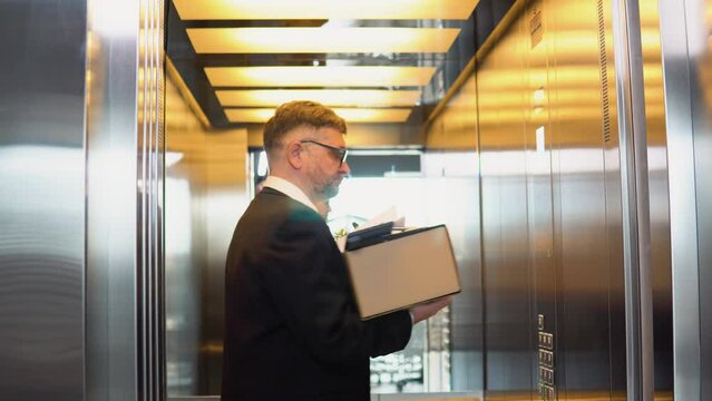 Sad dismissed senior worker with carton dismissal box getting into the elevator. Old man is retiring. Retirement concept. Dismissal from work, s3niorlife