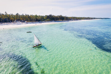 The tropical paradise of Zanzibar comes to life in this aerial view of fishing boats resting on the sandy beach at sunrise. The landscape features clear blue waters, lush palm trees, and a yacht in