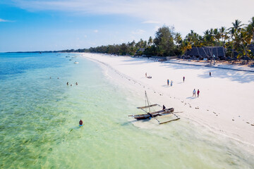 Take in the stunning scenery of Zanzibar's tropical coast from above, as fishing boats rest on the sandy beach at sunrise. The view from the top reveals clear blue waters, green palm trees, and even  - obrazy, fototapety, plakaty