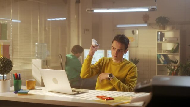 An angry and frustrated man in a yellow hoodie verifies the report, then angrily crumples a piece of paper and throws it away. A young creative team of managers is working in the background.