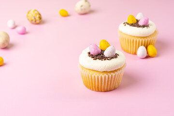 Two vanilla cupcakes decorated with cream, chocolate sprinkles and colorful glazed eggs on a blur on a pink background. Happy Easter Holiday.