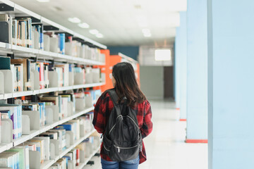 Smart student in casual style wearing backpack walking around the library while looking for a book, against bookshelf background. 