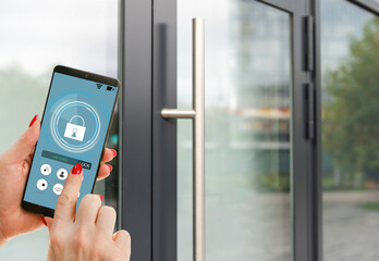 Female hand holding black mobile smart phone with smart home application on the screen. Blurred house on the background. For access to all of the controls of your house and caring of home security