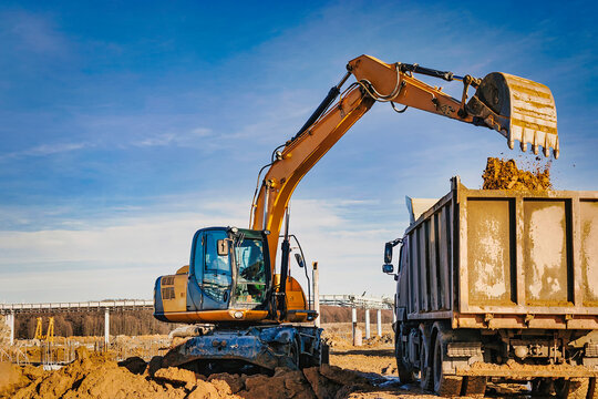 A wheeled excavator loads a dump truck with soil and sand. An excavator with a high-raised bucket against a cloudy sky View from the trench. Removal of soil from a construction site or quarry. - Powered by Adobe