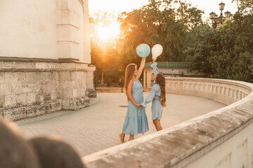 Obraz na płótnie Canvas Daughter mother run holding hands. In blue dresses with flowing long hair, they hold balloons in their hands against the backdrop of a sunset and a white building.