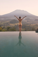Fototapeta na wymiar A woman stands on a pool overlooking the Agung volcano in Bali, watching the sunrise over calm water during her vacation.