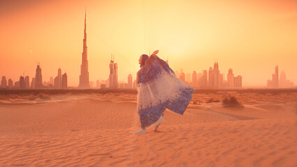 Woman in traditional arab dress stands on the mountains and rises her hand. Dubai city silhouette on the background
