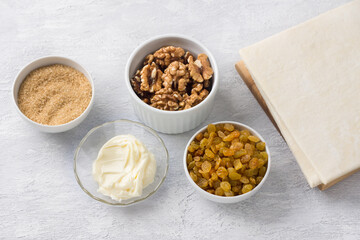 Ingredients for baking Easter cake kraffin: puff pastry, raisins, walnuts, soft butter, brown sugar on a light gray background, top view