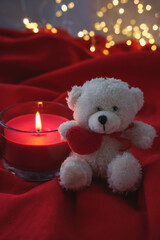 White teddy bear and burning candle on red fabric with bokeh background