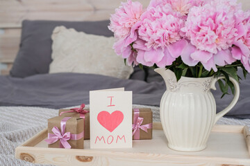 A vase of peonies and a card with the text I love mom on a tray on the bed.
