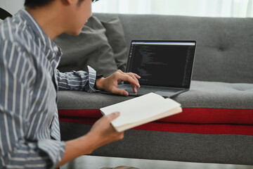 Male programmer sitting on floor in living room, working on laptop computer with programming code on screen