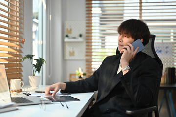 Asian male marketing sales manager consulting client, talking on mobile phone and using laptop pat working desk