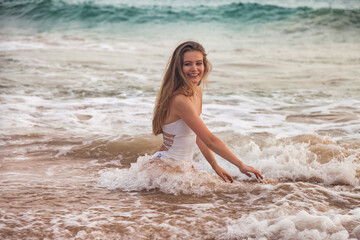 Fototapeta na wymiar Pretty young slim woman in white swimsuit posing in water at sea waves background, looking at camera. Relaxing and enjoyable vacation on tropics. Travel vacation holiday concept. Copy ad text space