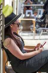 Elegant brunette girl wearing black hat and sunglasses sitting in the park with a cellphone