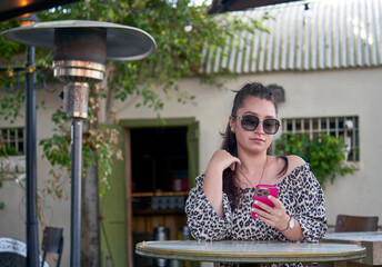 Beautiful brunette girl in sunglasses sitting in the open cafe in the park with a pink cellphone