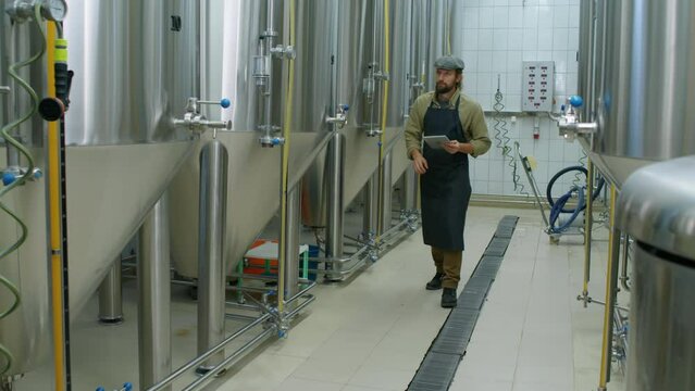 Full shot of young Caucasian male technologist in apron and cap walking along production facility at brewery, checking readings on gauges on bright beer tanks and completing report on tablet computer
