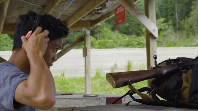 Side Portrait Of An Asian Man Puts On Protective Ear Muffs And Hold A Karabiner 98k At The Shooting Range. slowmo