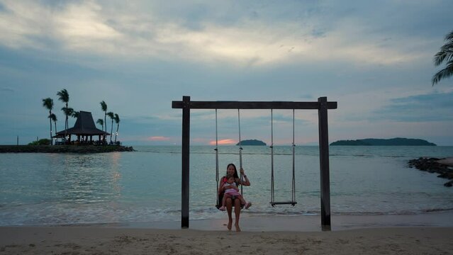 Mother with Little Daughter on Laps Swinging on Wooden Rope Swing At the Tanjung Aru Beach With Dramatic Colorful Sunset Sky in Background, Malaysia, Kota Kinabalu