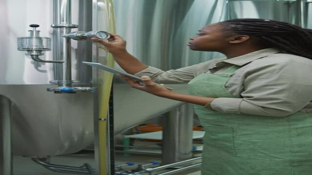 Medium vertical shot of African American female brewery worker in apron standing beside bright tank in fermentation room, taking readings from pressure gauge, filling out checklist on tablet computer